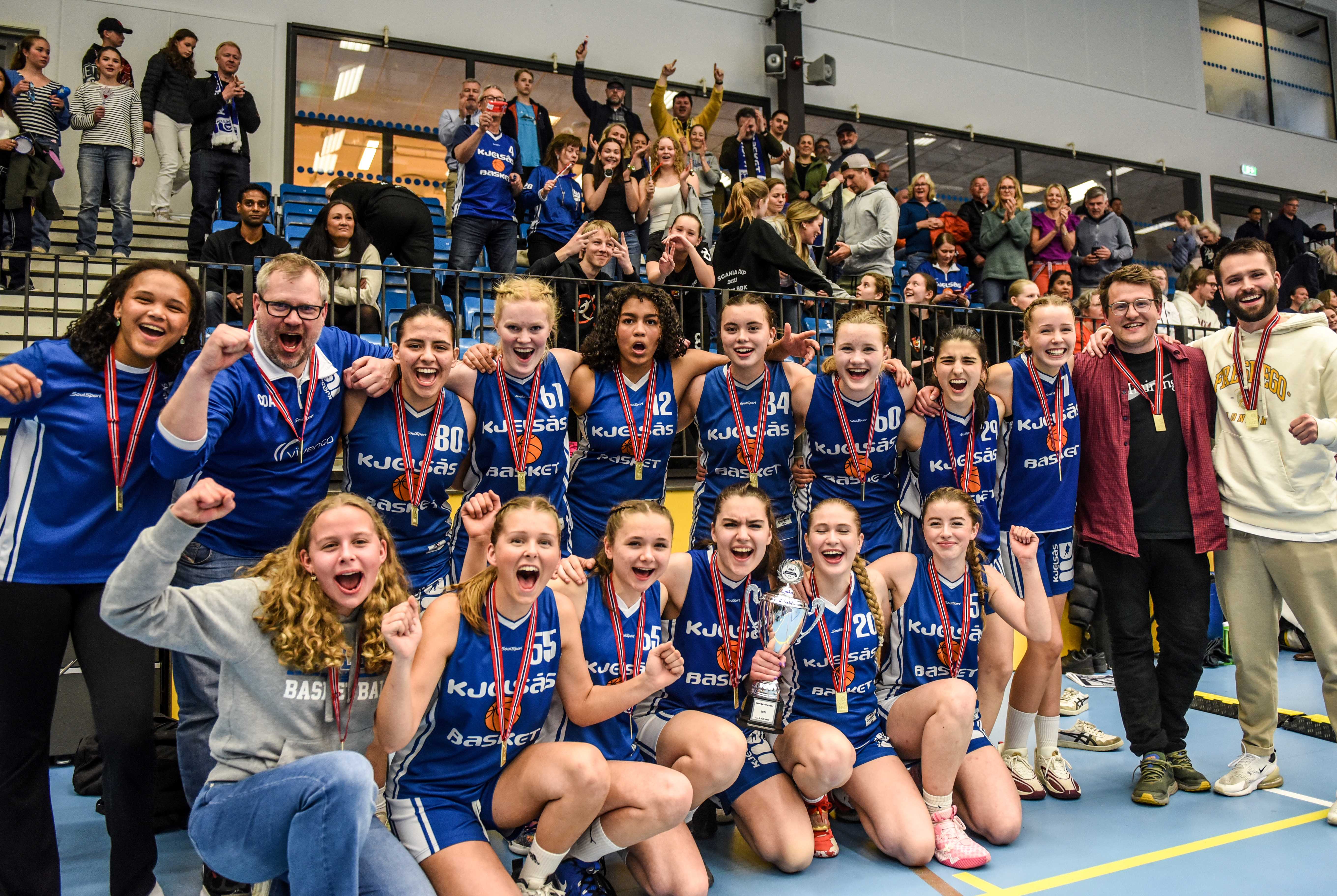 Kjelsås-jentene og fansen bak jublet for NM-gull etter finaleseier 61-54 over Ulriken Eagles. Bak fra venstre ser vi Nora Jazz Werenskiold Proctor, coach Omar Örn Sigmarsson, Emina Mutevelic, Cora Fiskevoll, Andrea Fergem Faal, Sofie Rime Jobling, Oda Marie Tveiten, Sofija Jozin, Johanna Kolseth Kiland, Jørn Nordal og Peder Kletten. Foran fra venstre Celine Mannsåker, Erle Svendsen Hektoen, Kari Lamvik Horn, Sara Ivankovic, Rakel Sif Omarsdottir og Inga Hesthammer. 
FOTO: ERIK BERGLUND 